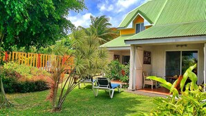 A house with a green roof, a covered porch with chairs, and a garden with various plants and a wooden fence.