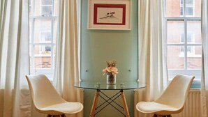 Ein Zimmer mit Glastisch und zwei weißen Stühlen, einem gerahmten Bild an der Wand und einem Fenster mit Vorhängen.