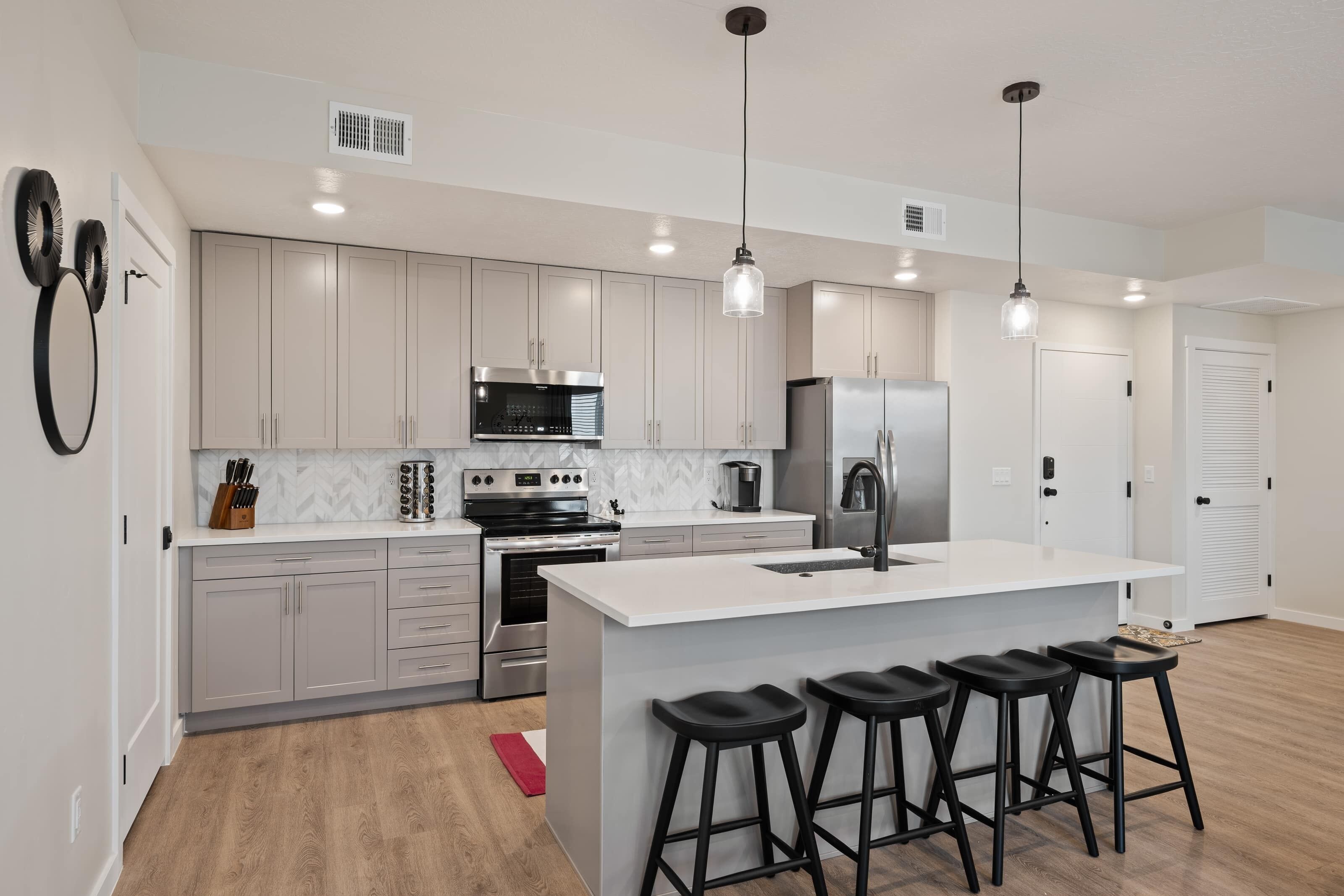 A modern kitchen with white cabinets, stainless steel appliances, a central island with bar stools, pendant lighting, and a red rug on the floor.