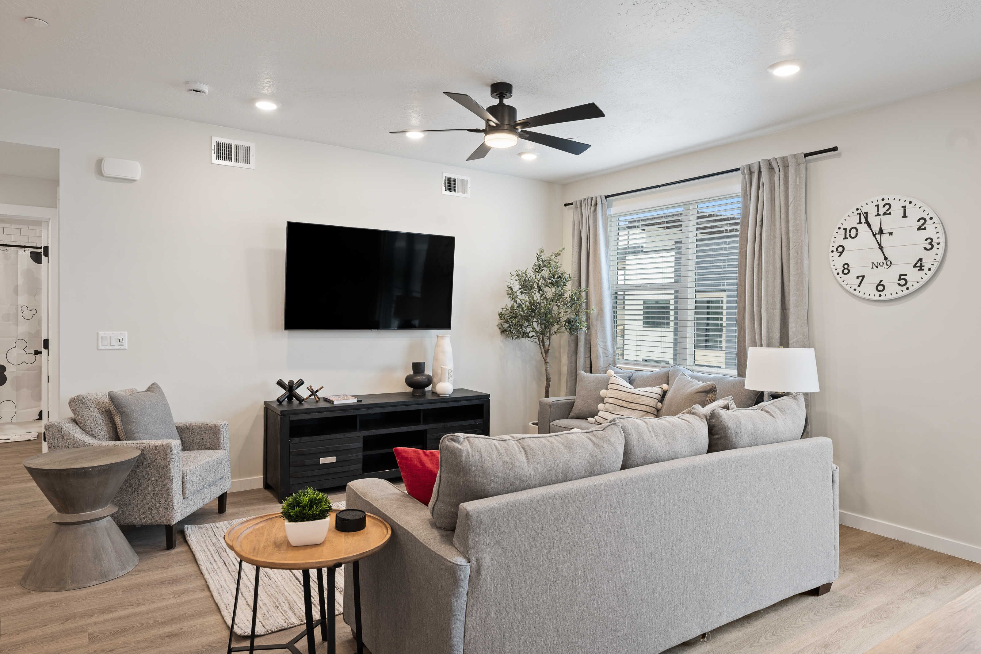 A modern living room with a grey sofa, a black entertainment unit, a flat-screen TV, a ceiling fan, and a large wall clock.