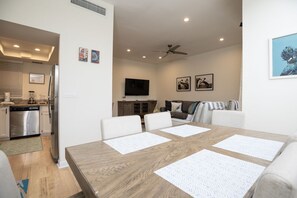 Een moderne keuken met roestvrijstalen apparatuur, een eetgedeelte met een houten tafel en witte placemats, een woonkamer met een bank en een televisie, en kunstwerken aan de muur.