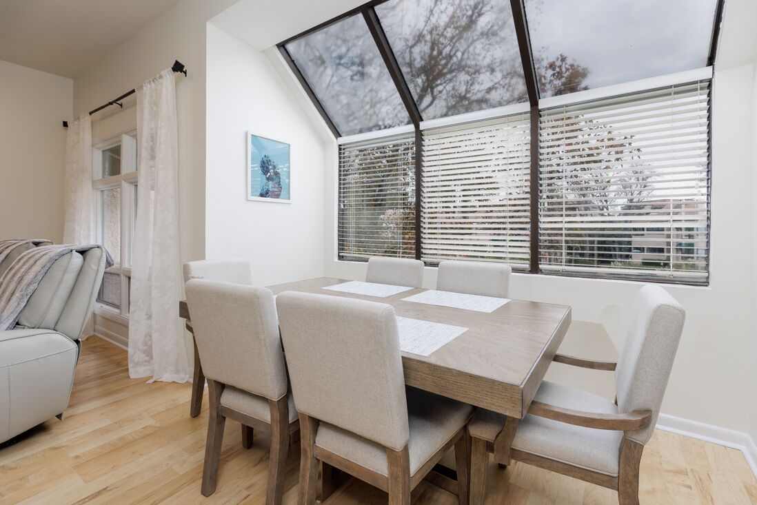 Een eetruimte met een houten tafel en zes stoelen, een bank en een raam met zonwering.