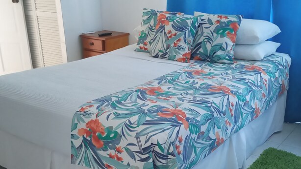 Ein ordentlich bezogenes Bett mit floralem Überwurf und Kissen, ein kleiner Nachttisch aus Holz und ein grüner Teppich.