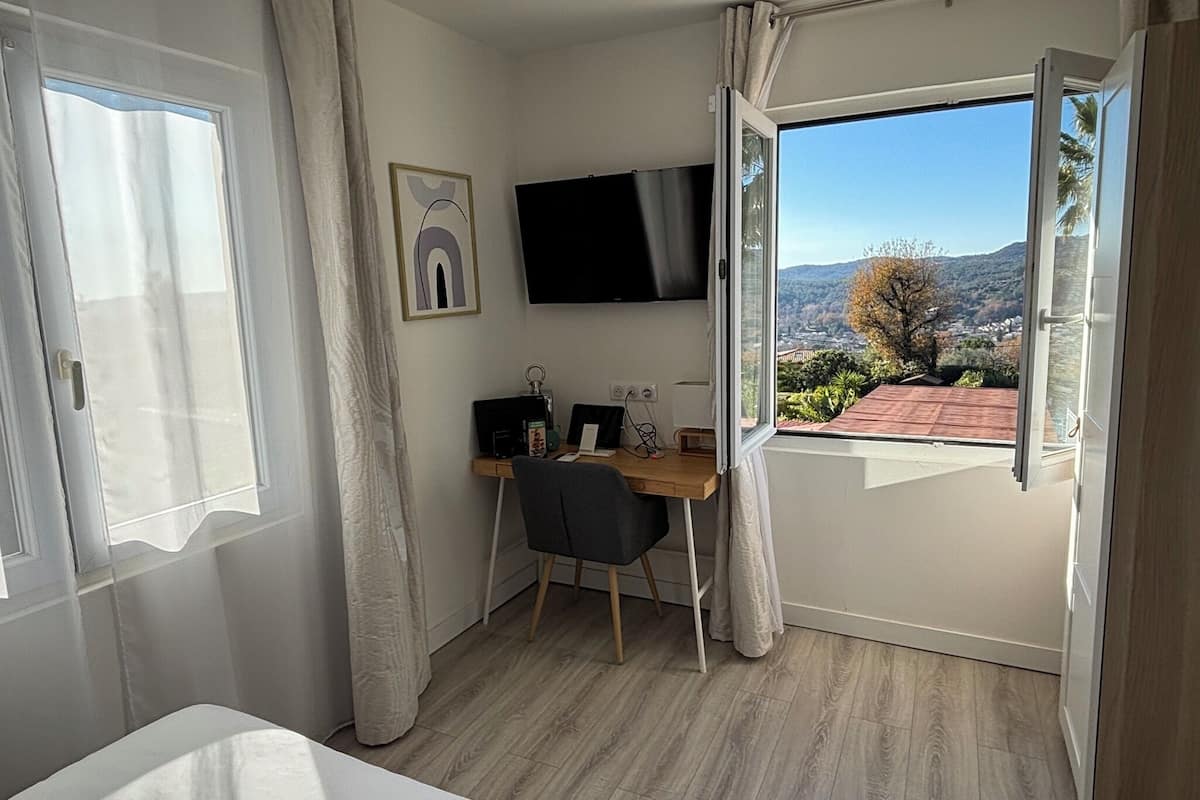 A bedroom with a bed, a desk with a chair, a TV, and a window with a view of a garden and hills.