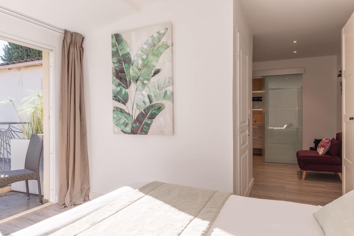 A bedroom with a bed, a large window with curtains, a plant, and a sliding door leading to a balcony.