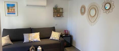A modern living room with a dark grey sofa, patterned cushions, a small black side table with a decorative item, and a wall-mounted air conditioner.