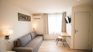Una habitación compacta con sofá gris, un escritorio pequeño con silla, televisor de pared y una puerta.