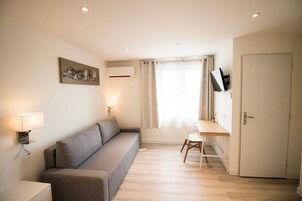 Una habitación compacta con sofá gris, un escritorio pequeño con silla, televisor de pared y una puerta.