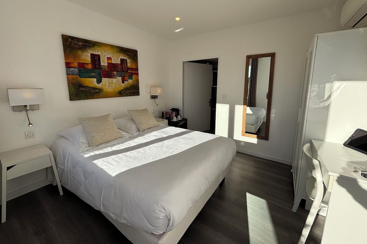 A modern hotel room with a large bed, bedside tables, a desk, a chair, and a painting on the wall.