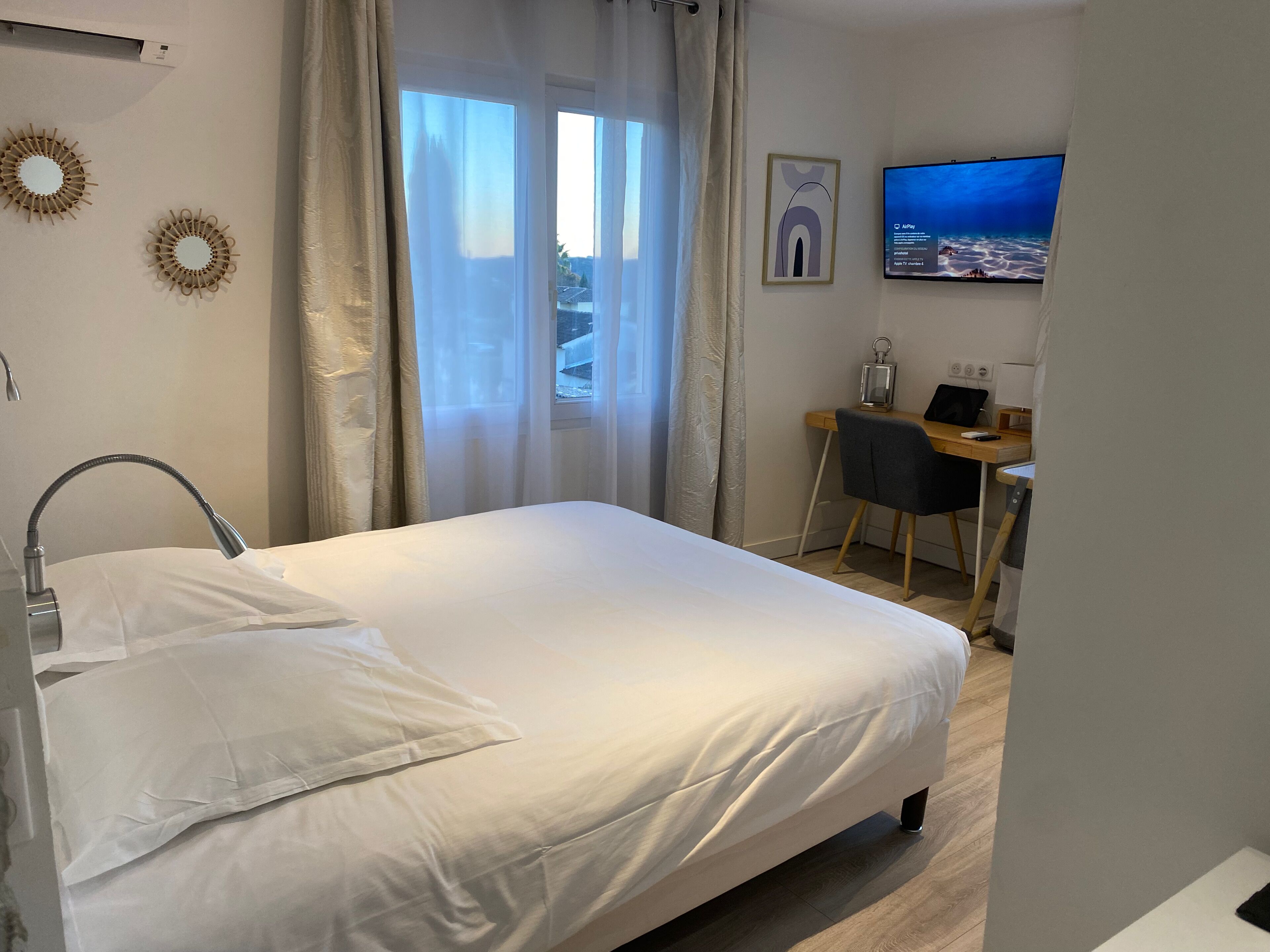 A neatly made bed with white linens, a desk with a chair, a television mounted on the wall, and a window with sheer curtains.