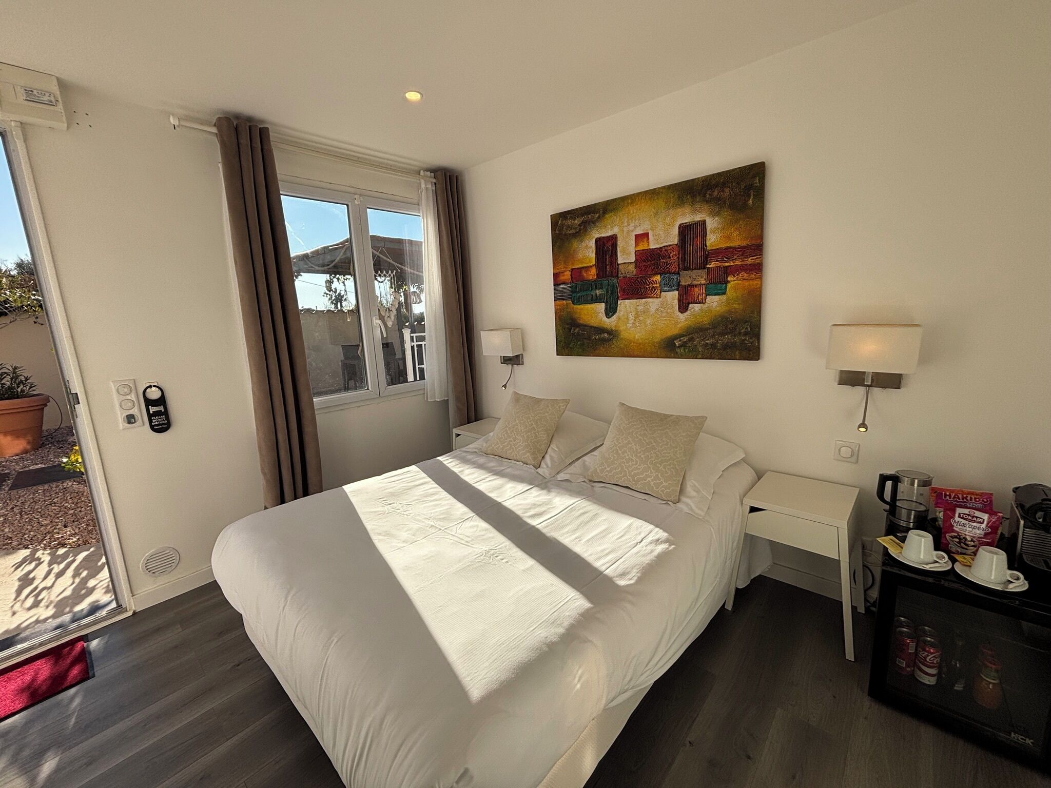 Habitación de hotel con una cama grande, un cuadro en la pared, una ventana con cortinas, una mesita de noche con una lámpara y una cafetera.