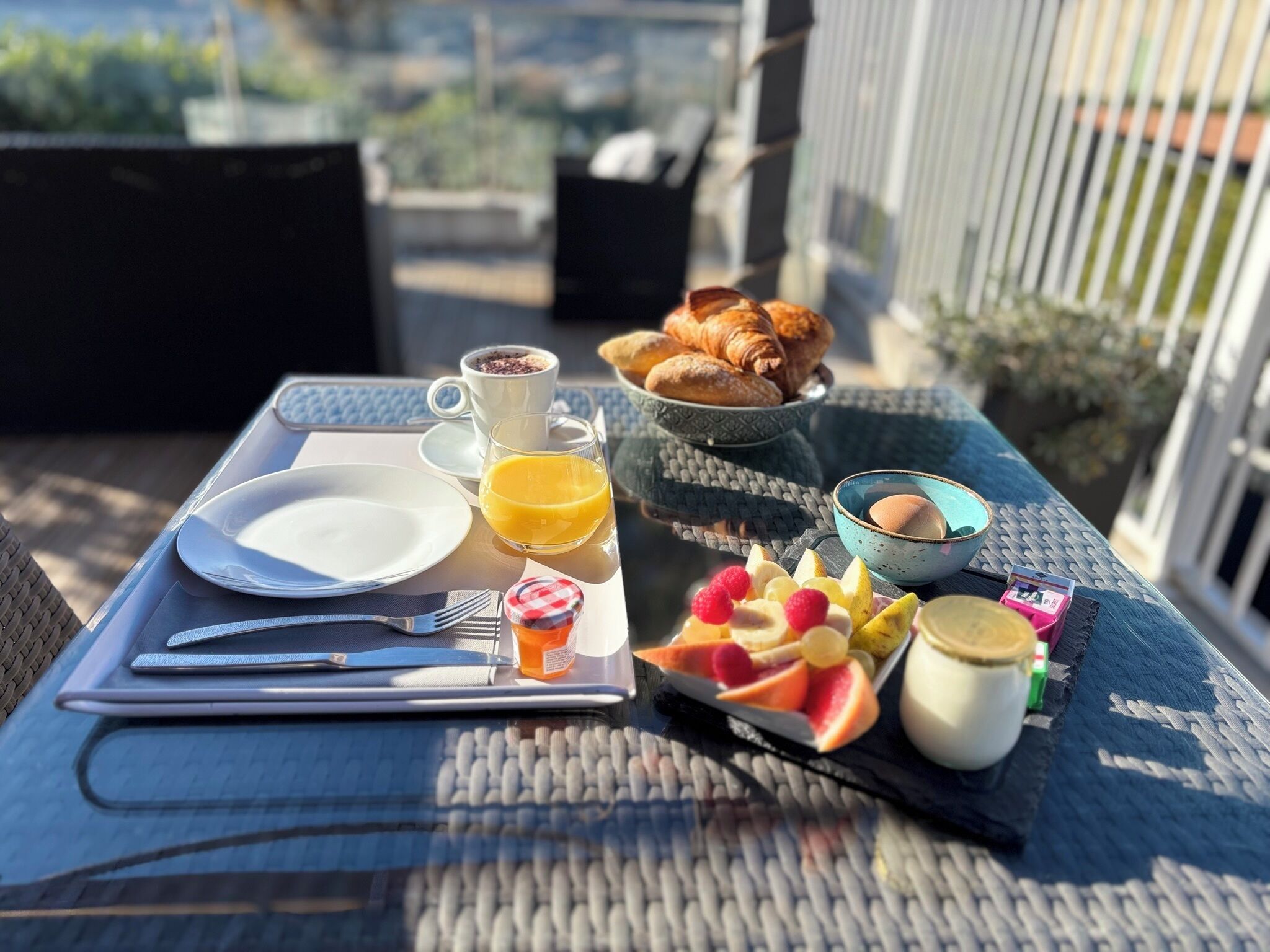 Una mesa de desayuno con croissants, café, zumo de naranja y fruta en un balcón.