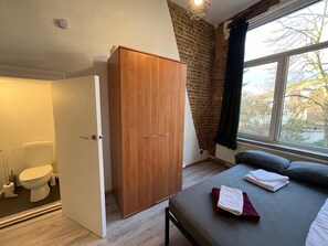 A bedroom with a wooden wardrobe, a bed with pillows, a window with a view of trees, and a bathroom with a toilet and shower.