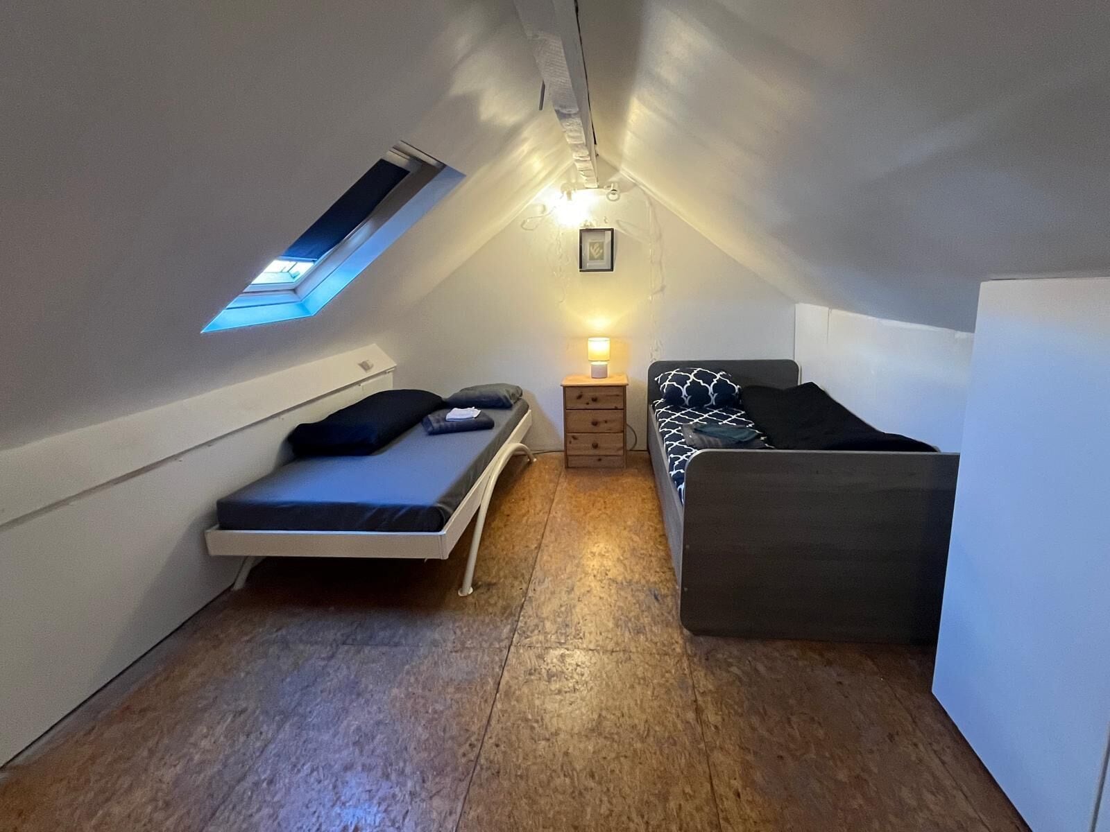 Una habitación pequeña y estrecha, con techo inclinado, una cama individual, una mesita de noche con lámpara y un ventanal.