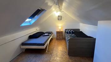 Una habitación pequeña y estrecha, con techo inclinado, una cama individual, una mesita de noche con lámpara y un ventanal.