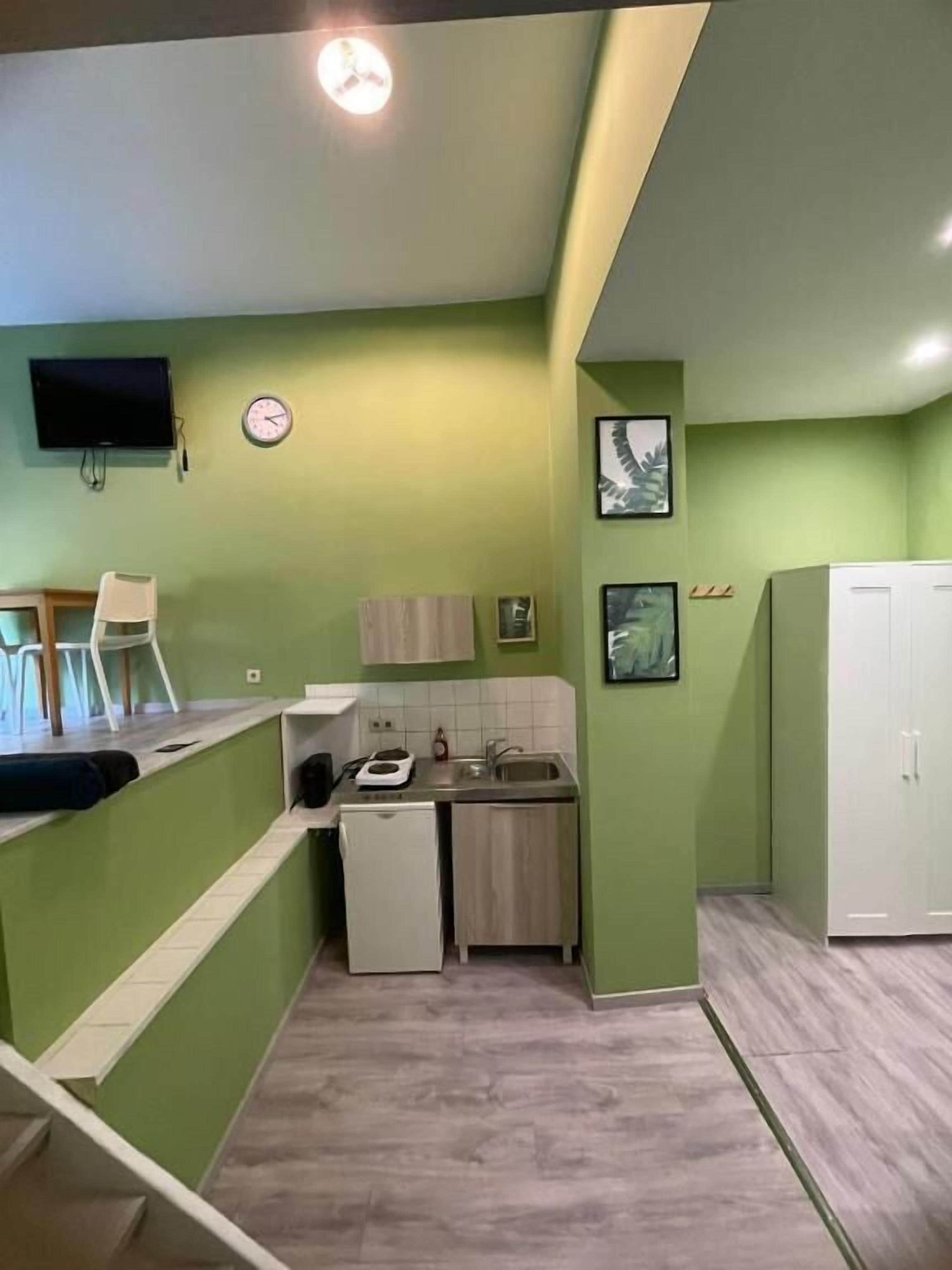 Een kleine keuken met een spoelbak, een kleine koelkast en een magnetron. Er hangt een klok aan de muur en erboven een televisie.