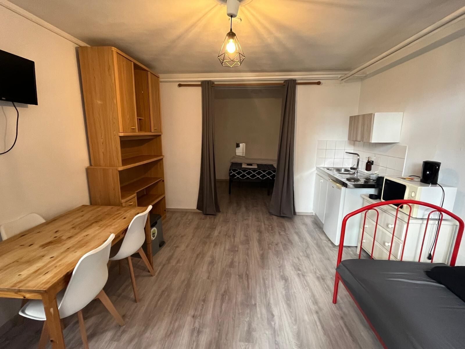 Een compacte kamer met een houten eettafel, een bed met rood metalen frame, een kitchenette met witte kastjes en een plafondlamp.