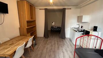 Een compacte kamer met een houten eettafel, een bed met rood metalen frame, een kitchenette met witte kastjes en een plafondlamp.