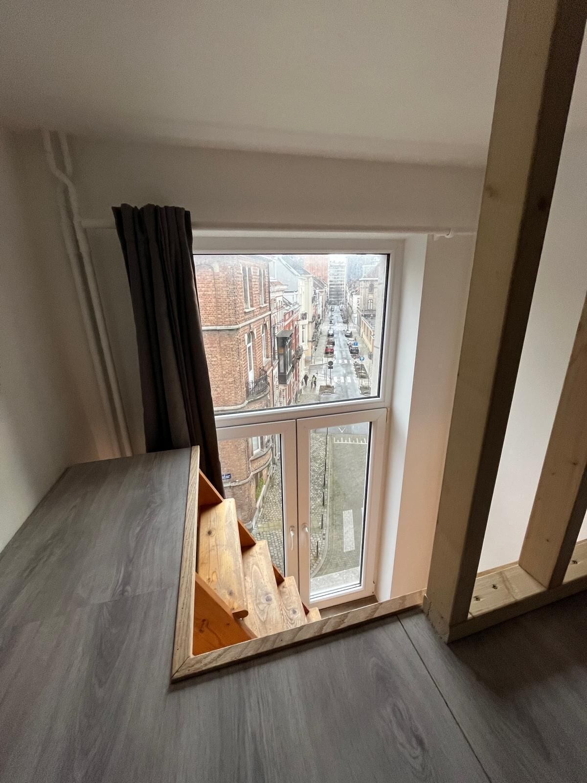 Ein Zimmer mit großem Fenster, einer hölzernen Bank und Blick auf eine Stadtstraße.