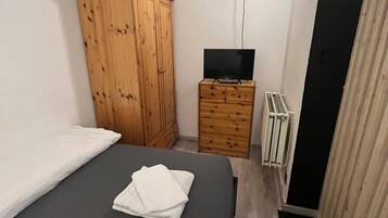 Et lille, kompakt værelse med en enkelt seng, et træskab, en lille kommode, en radiator og et fjernsyn.