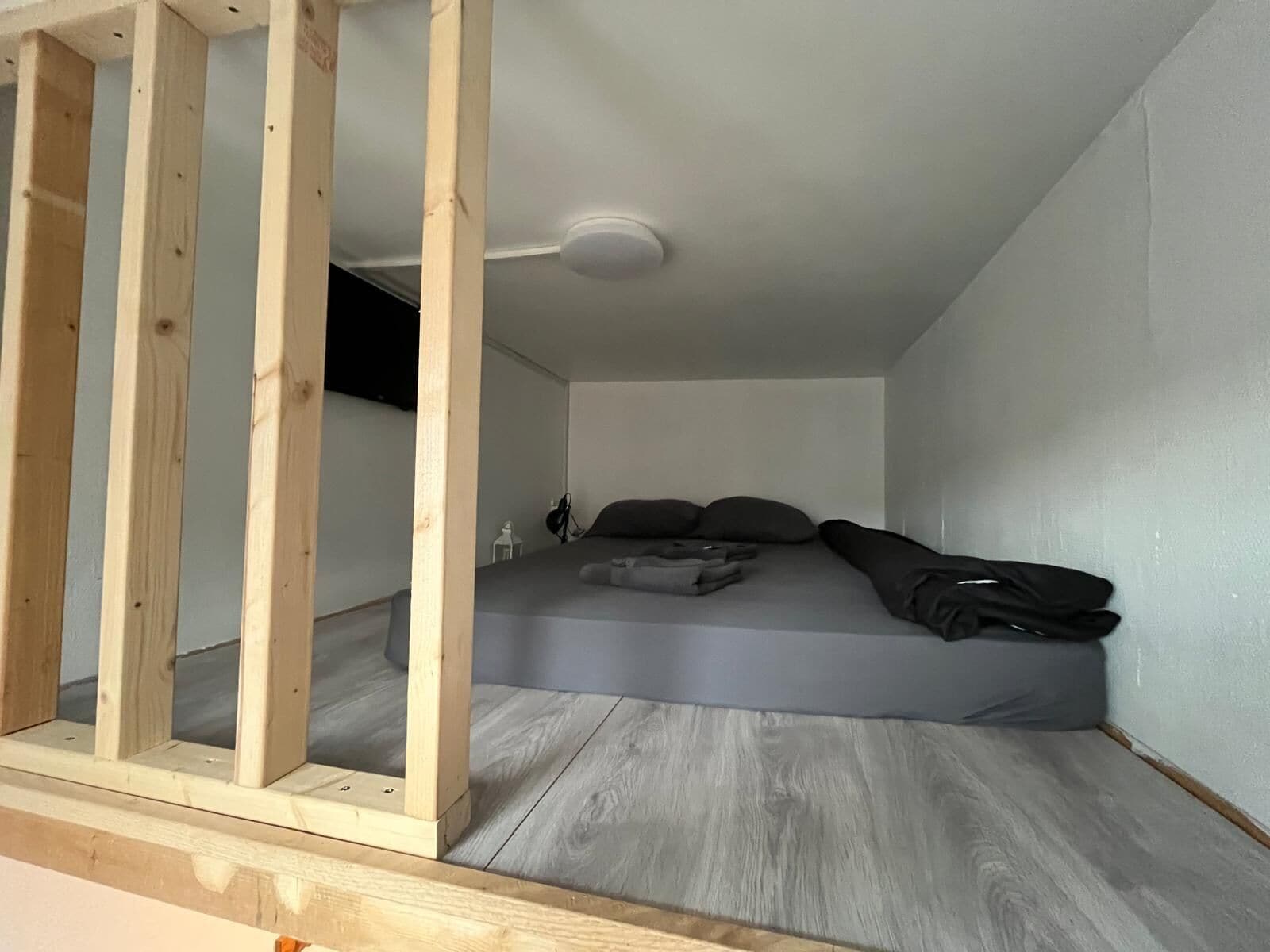 Una camera piccola con un letto singolo, materasso grigio, poggiatesta in legno e ventilatore a soffitto.