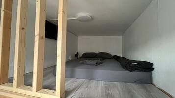 Una camera piccola con un letto singolo, materasso grigio, poggiatesta in legno e ventilatore a soffitto.