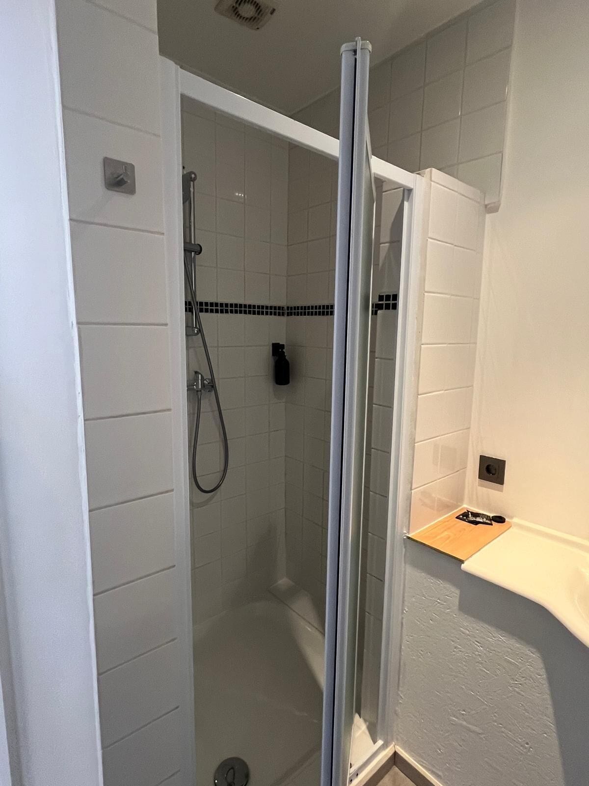 A modern bathroom with a glass-enclosed shower, white tiled walls, and a wooden shelf.