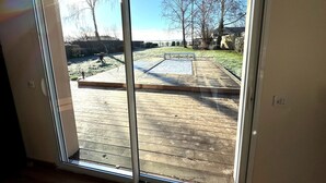 A view through a sliding glass door to a wooden deck with a metal bench and a barbecue grill in the yard. - Charming Family House with Garden and Heated Swimming Pool 1 hour from Paris (Méré)