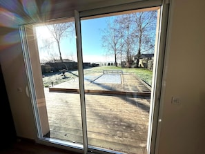 Vista a través de una puerta corrediza de vidrio a una terraza de madera con un banco de metal y una parrilla en el jardín.