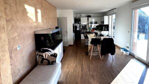A modern living room with a sofa, a dining area with a table and chairs, and a kitchen in the background. - Charming Family House with Garden and Heated Swimming Pool 1 hour from Paris (Méré)