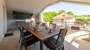 Un espace extérieur aménagé pour les repas, avec une table et des chaises en bois, un barbecue et une vue sur une maison et des arbres.