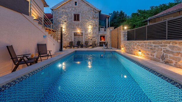 Une piscine aux murs en pierre, un bâtiment en pierre en arrière-plan et des sièges à l’extérieur.