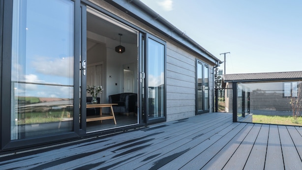 A modern house with large sliding glass doors leading to an interior space, a wooden deck, and a clear sky. - Bluebell Lodge (Dartmouth)