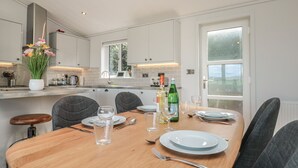 A modern kitchen with white cabinets, a wooden dining table set for four, and a view of the outdoors through a large window. - Bluebell Lodge (Dartmouth)