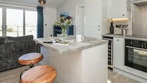 A modern kitchen with white cabinets, a built-in oven, and a dining area with a bar stool and a magazine. - Bluebell Lodge (Dartmouth)