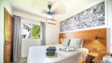 A hotel room with a bed, a ceiling fan, an air conditioning unit, a window with curtains, and a wall with a leaf pattern.
