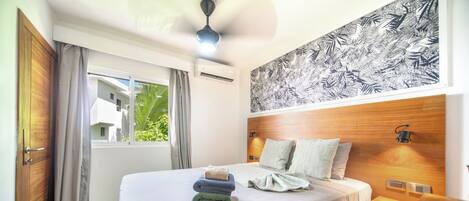 A hotel room with a bed, a ceiling fan, an air conditioning unit, a window with curtains, and a wall with a leaf pattern.