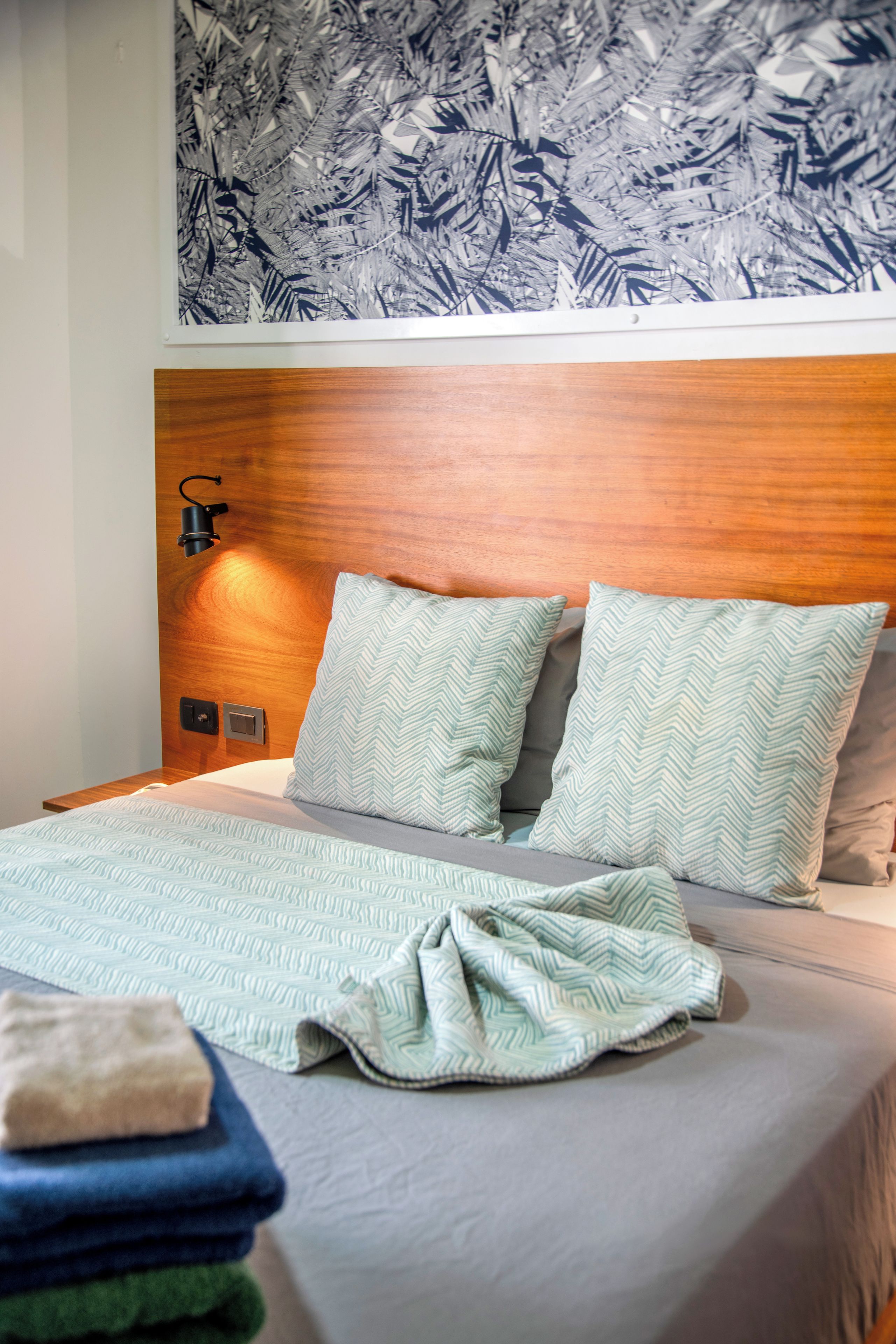 Een net opgemaakt bed met een bedspreide en kussens met een patroon, een houten hoofdbord, een wandlamp en een decoratieve wand boven het bed.
