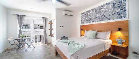 A modern hotel room with a bed, a ceiling fan, a wall-mounted air conditioner, a dining area with a table and chairs, and a balcony with curtains.