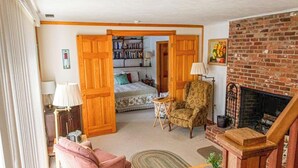 Un salon chaleureux avec une cheminée, un fauteuil confortable, un canapé et un lit à l’arrière-plan.