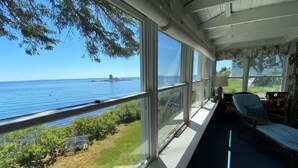 A room with large windows overlooking a body of water, a chair, and a small table. - Experience coastal charm at Edge Of The Sea, a cozy 3-bedroom cabin in Boothbay, ME. (Boothbay)