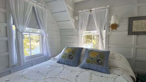 A bedroom with a bed, two windows with curtains, a wall-mounted lamp, and a framed picture. - Experience coastal charm at Edge Of The Sea, a cozy 3-bedroom cabin in Boothbay, ME. (Boothbay)