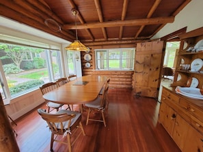 Un coin repas rustique en bois, avec une grande table et des chaises, un luminaire suspendu et une vue sur l’extérieur grâce à de grandes fenêtres.