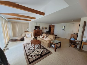 Un salon spacieux avec un plafond haut, des poutres apparentes, un canapé beige, une table basse et un tapis à motifs.