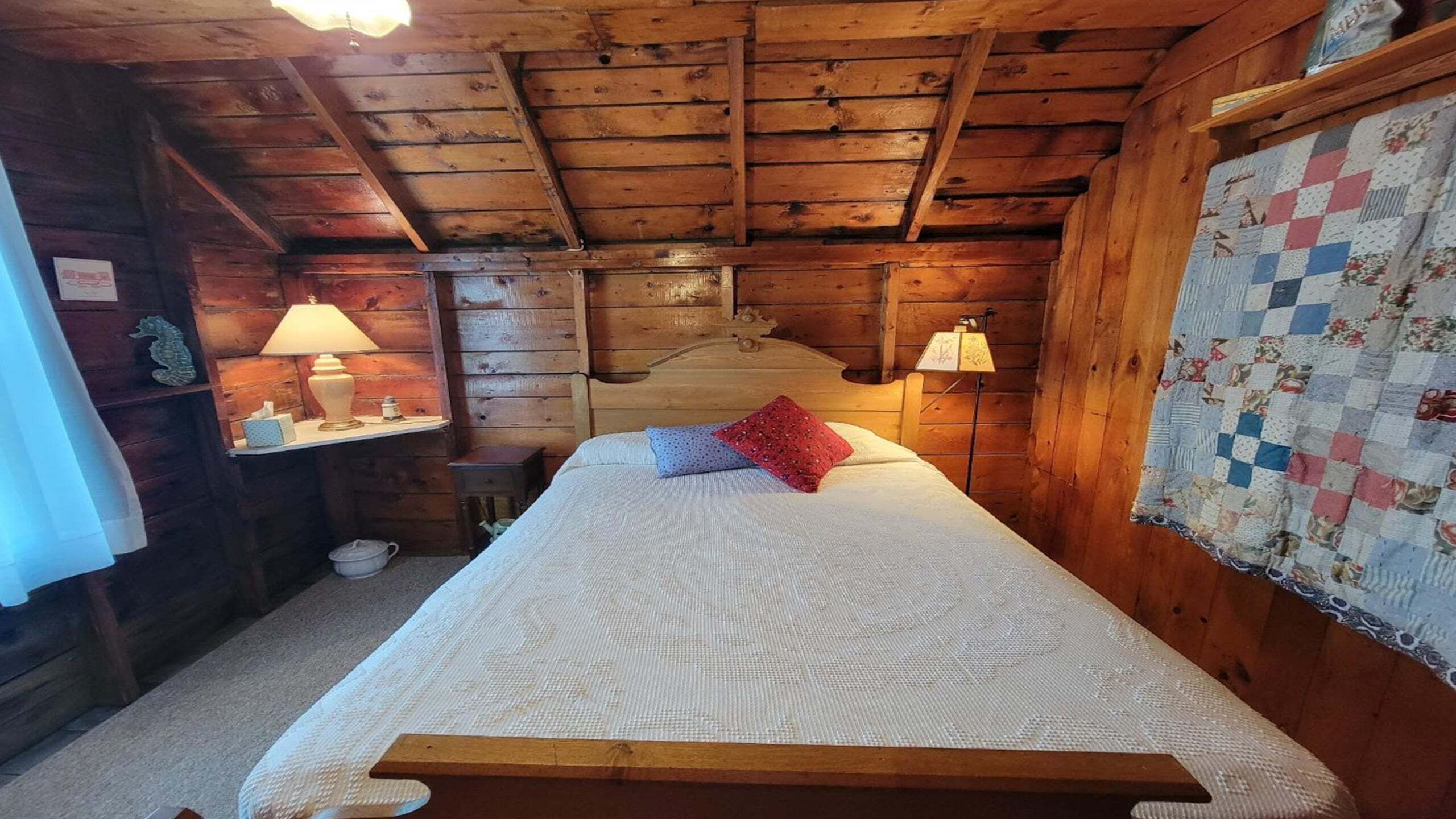Et hyggeligt værelse i en træhytte med seng, to sengeborde med lamper, et tæppe og en trægavl.