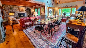 A spacious living area with a stone fireplace, wooden beams, a dining table with chairs, and a large window providing natural light. - Experience coastal charm at Tidewatch in Boothbay, a spacious 3-bedroom home near the ocean. (Boothbay)