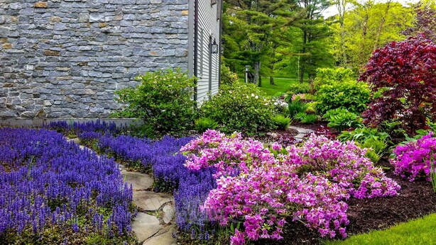 A stone building with a garden featuring purple and pink flowers, a stone pathway, and a metal fence. - Experience coastal charm at Tidewatch in Boothbay, a spacious 3-bedroom home near the ocean. (Boothbay)