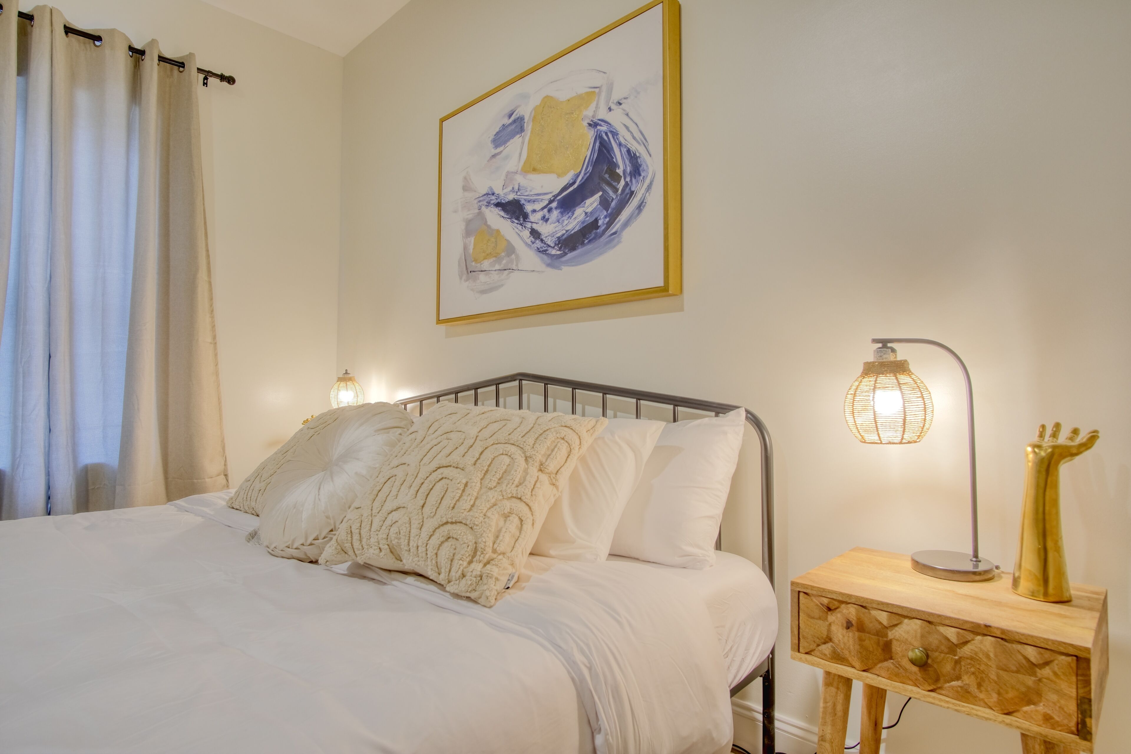 A neatly made bed with white linens and decorative pillows, a bedside table with a lamp, and a framed abstract painting on the wall.