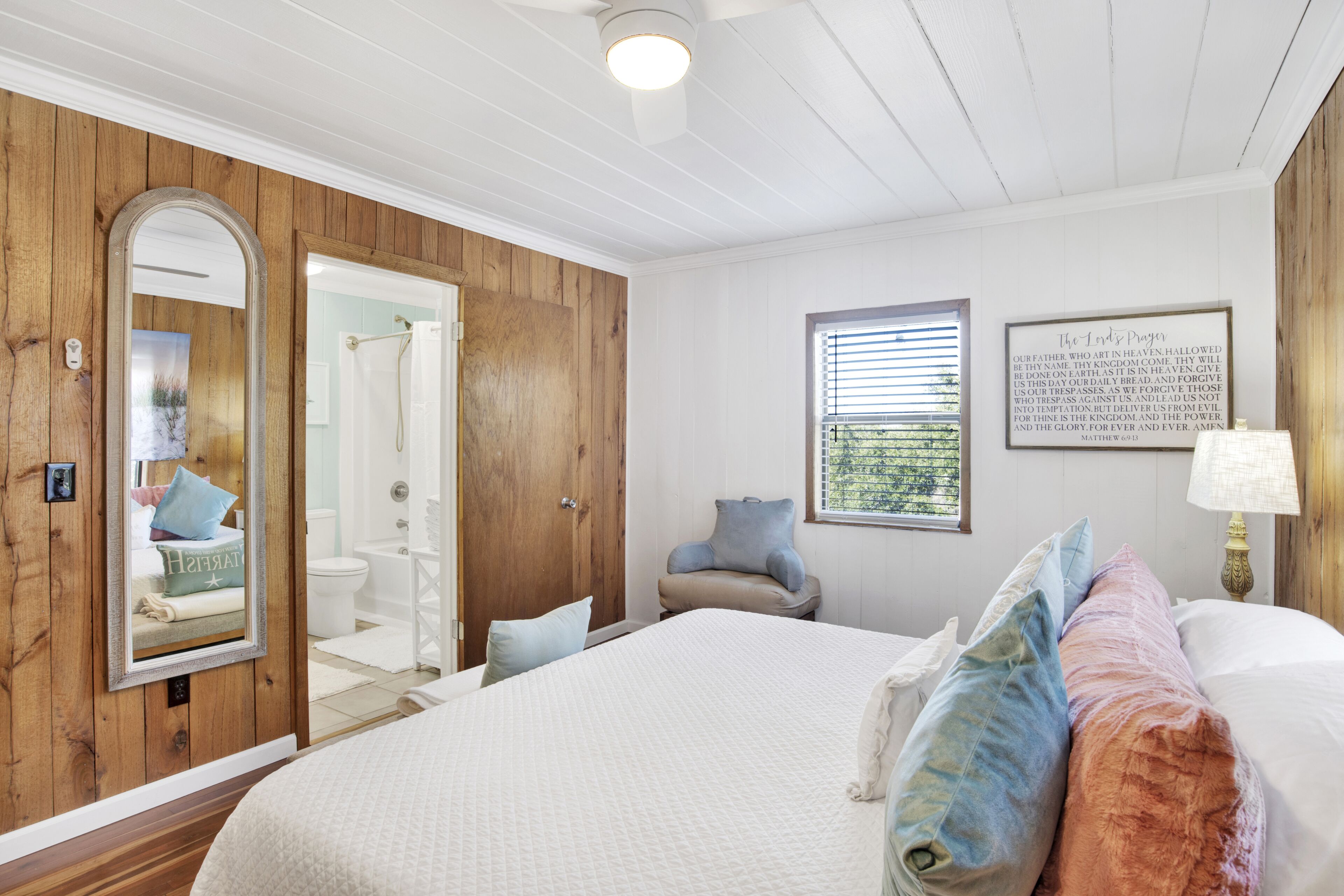 Een slaapkamer met een groot bed, houten paneelwanden, een spiegel, een raam met zonwering en een lijst met een citaat aan de muur.