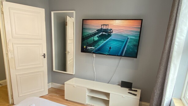 Quarto de hotel com uma cama, televisão fixada na parede, mesa consola branca e uma porta.